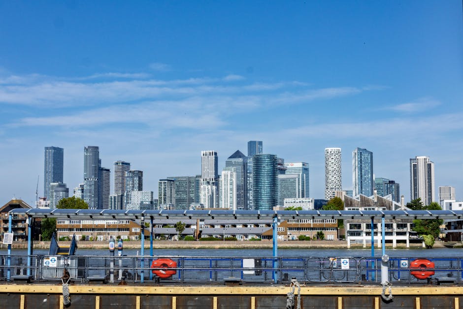 A panoramic view of Canary Wharf skyline featuring modern glass skyscrapers, with a clear blue sky overhead. In the foreground, a riverfront with a dock structure, including lifebuoys and safety raili