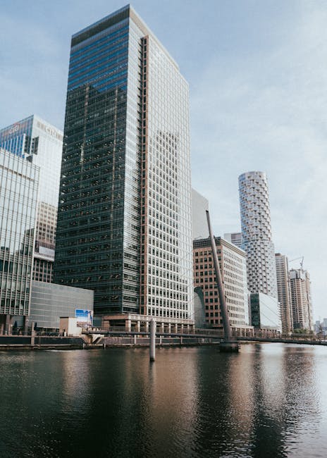 A panoramic view of Canary Wharf skyline featuring modern glass skyscrapers, with a clear blue sky overhead. In the foreground, a riverfront with a dock structure, including lifebuoys and safety raili