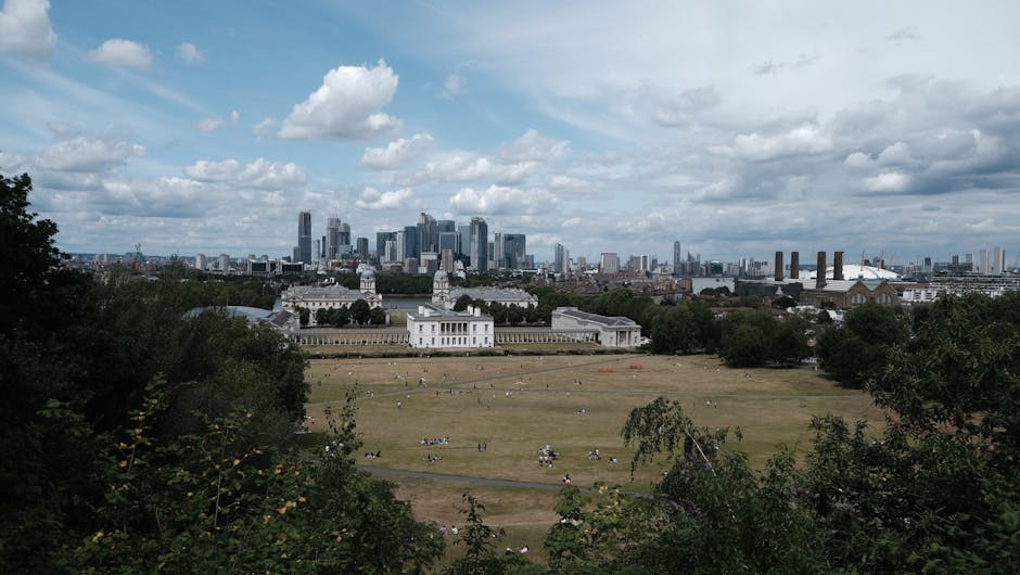 A wide view of the Jubilee Park communal area, showing a large green open space with neatly maintained lawns and scattered visitors. In the background, a modern cityscape features tall glass skyscrape