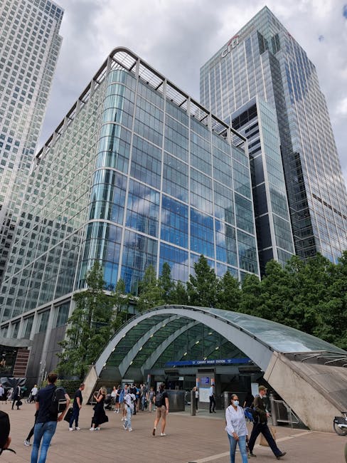 Exterior view of modern office buildings in Canary Wharf, featuring glass curtain walls and steel framing, with reflections of the blue sky. The buildings have sleek, curved and rectangular surfaces, 